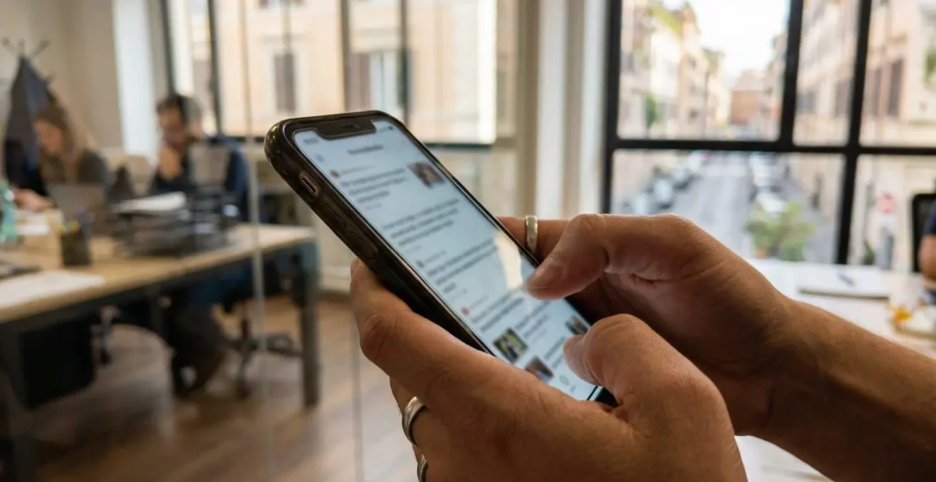 Dettaglio di mani che scorrono su uno smartphone con lo sfondo sfuocato di una finestra d'ufficio